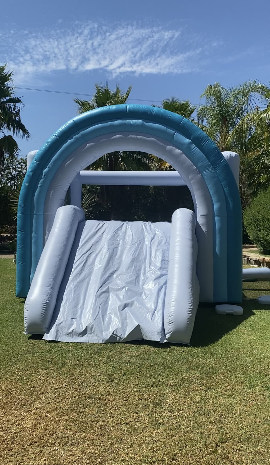 Insuflável azul e branco com escorrega montado num jardim com palmeiras