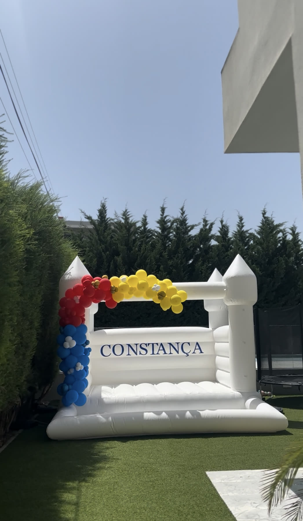 Castelo insuflável branco personalizado com nome e arco de balões coloridos num jardim